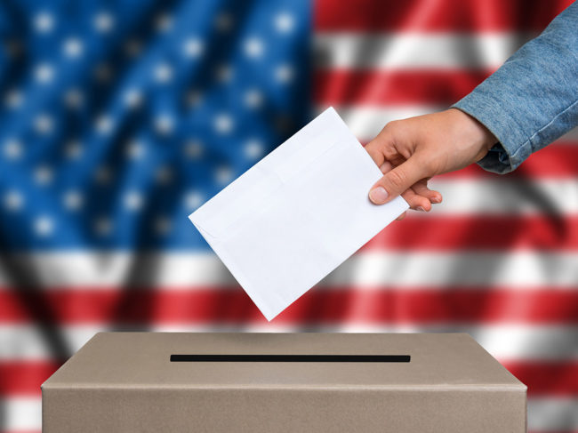 Hand holding ballot over box, US flag backdrop