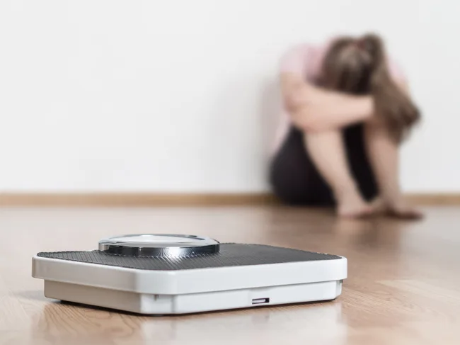 Photo of scale with woman sitting on floor holding head and arms on knees