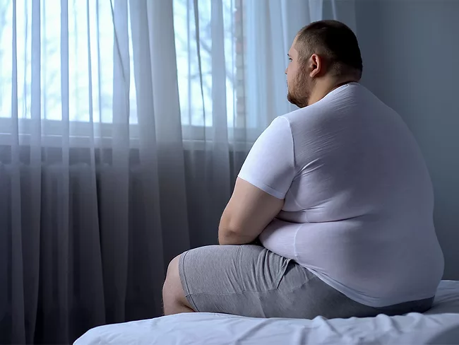 Overweight man sitting on bed facing the window